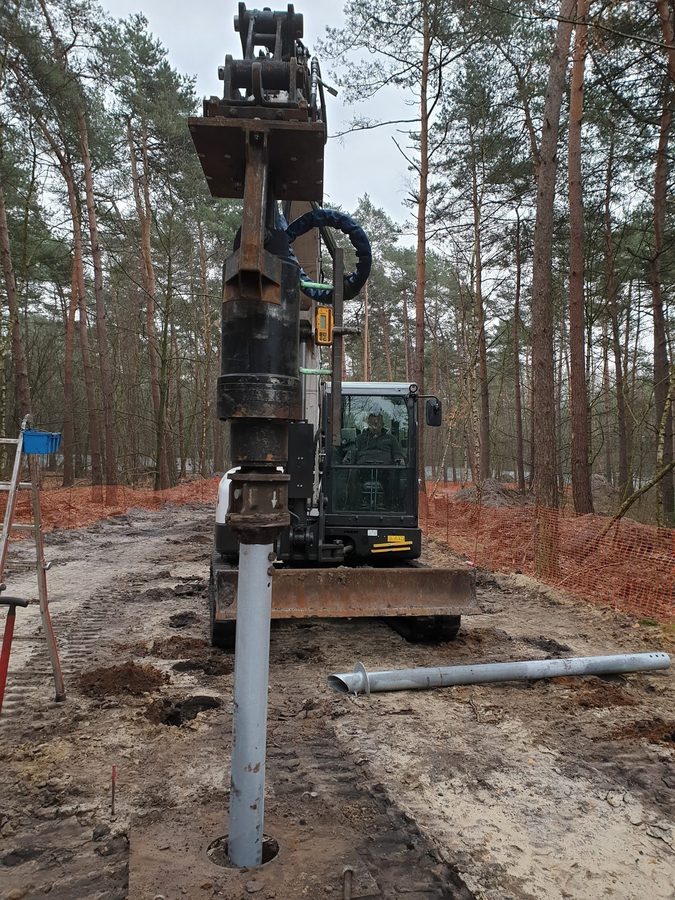 Grävmaskin installerar varmförzinkad skruvpåle bland tallar i Boslands skog, Belgien